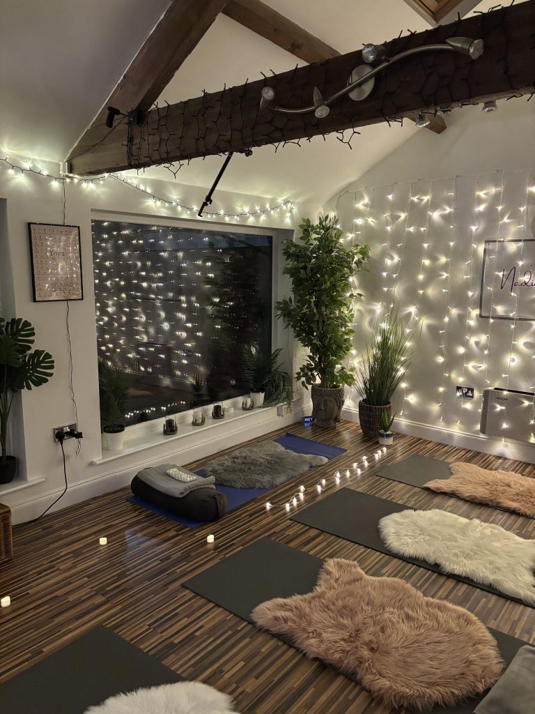 Yoga by Nadine Hill studio, image shows fairy lights in the background and yoga mats, blankets and sheepskin rugs set up on the wood panelled floor.