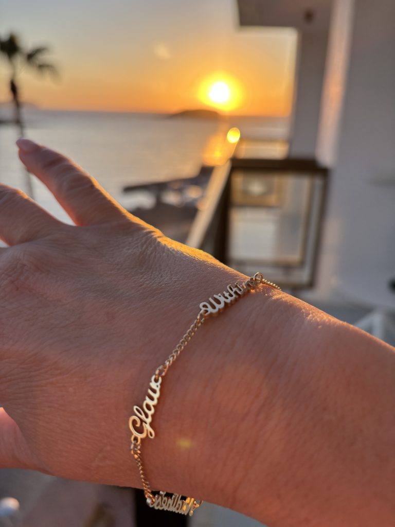 Image of a hand wearing a gold bracelet that says the words: Nadine's Glow Within on it, in front of a sunrise over sea