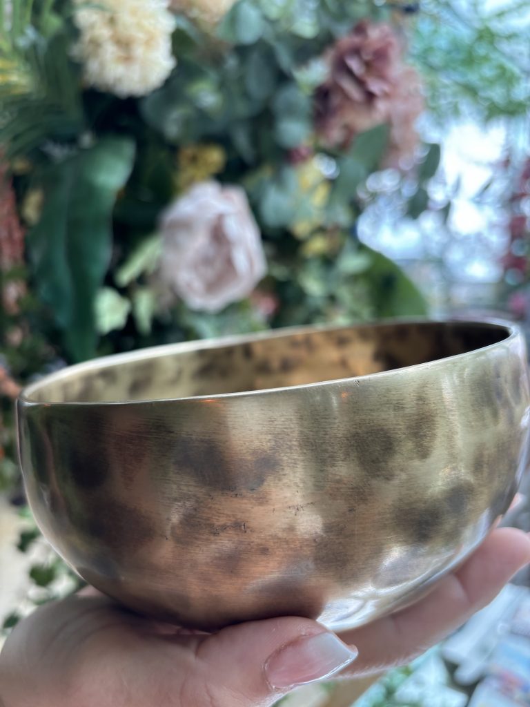 a white hand palm up, holding a Himalayan singing bowl and flowers in the background.