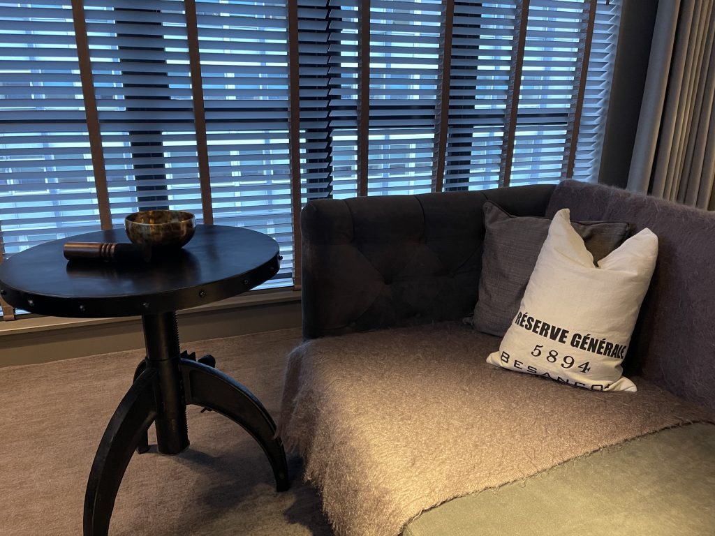 Executive setting. Image of a smart structured sofa next to a side table which contains a brass coloured Himalayan singing bowl and wand, in front of a large window covered with a dark Venetian blind.